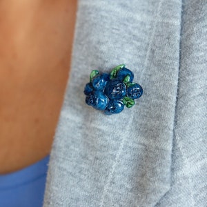 Könnte beinhalten: Eine blaue und grüne Blaubeer-Brosche. Die Brosche ist aus Polymer Clay gefertigt und hat die Form einer Blaubeertraube.