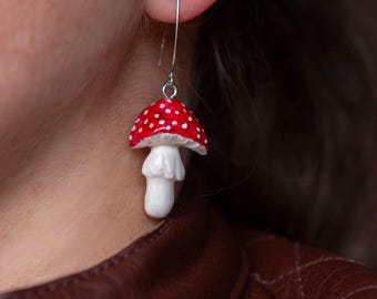 Op de afbeelding: Een enkele paddenstoeloorbel met een rode hoed en witte stippen. De paddenstoel heeft een witte steel en is opgehangen aan een zilverkleurige haak. De oorbel is een grillig accessoire.