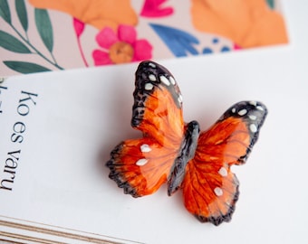 May include: A ceramic butterfly brooch with orange and black wings and white spots. The butterfly is sitting on a white book with the text "Kó es varu" printed on the page.