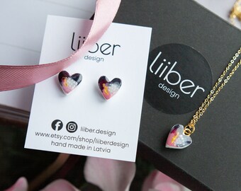 Puede incluir: Un conjunto de pendientes en forma de corazón y un collar con un colgante en forma de corazón. Las joyas presentan un diseño abstracto rosa y blanco con detalles dorados. Las joyas están empaquetadas en una caja negra con una etiqueta blanca que dice "liiber design".