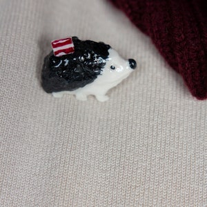 May include: A small white and black ceramic hedgehog figurine with a red and white striped hat. The figurine is sitting on a beige knitted fabric.