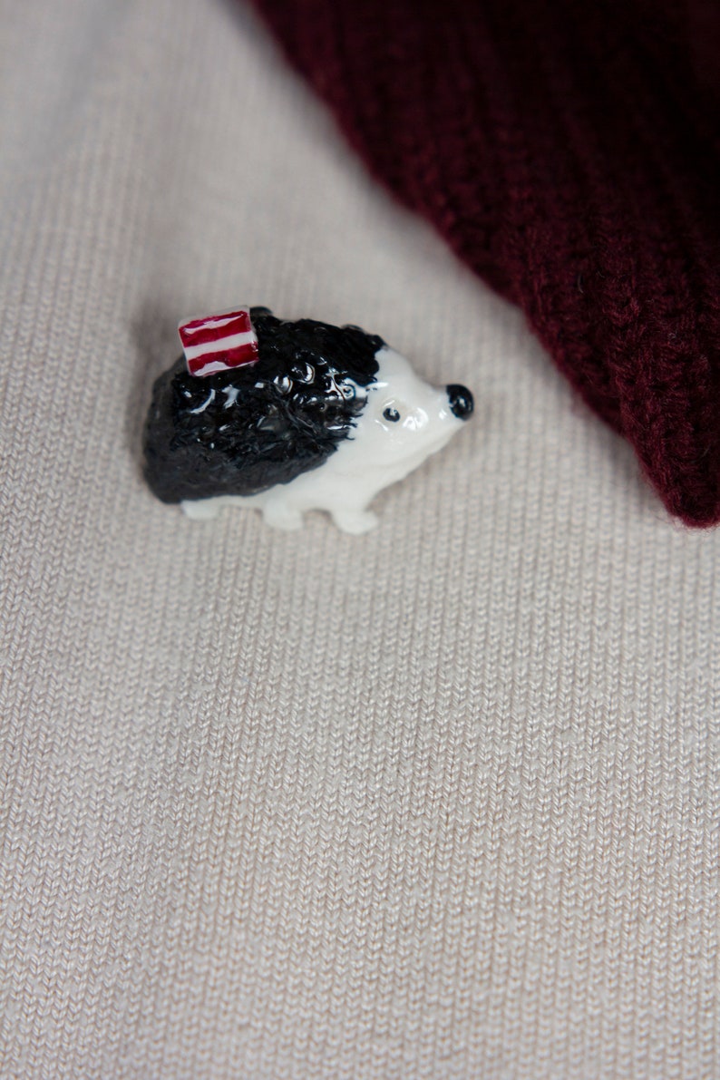 May include: A small white and black ceramic hedgehog figurine with a red and white striped hat. The figurine is sitting on a beige knitted fabric.