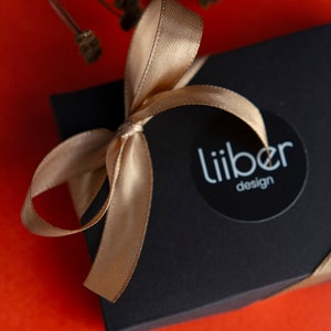 May include: A black gift box with a gold ribbon tied in a bow. The box has a white circle sticker with the text "Liiber design" on it.