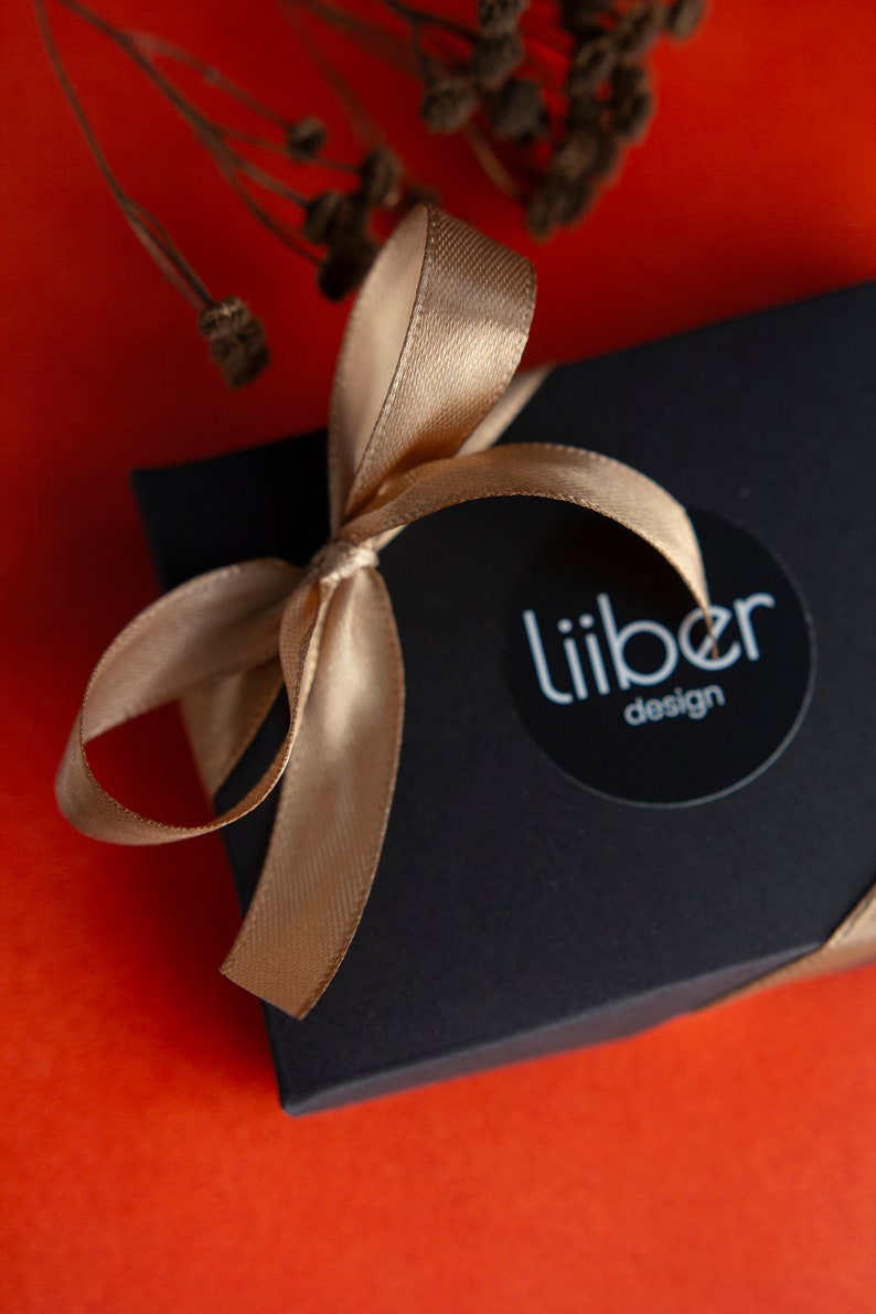 May include: A black gift box with a gold ribbon tied in a bow. The box has a white circle sticker with the text "Liiber design" on it.