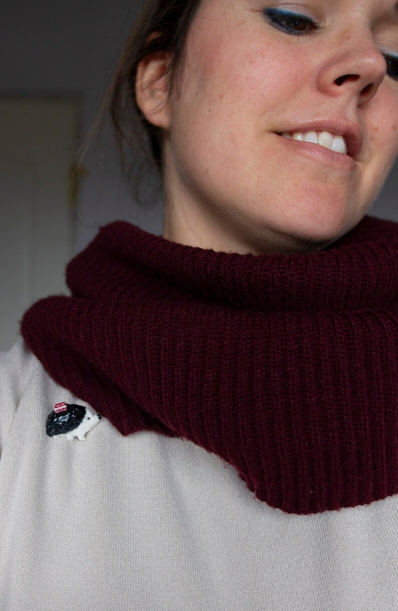 May include: A woman wearing a maroon knitted cowl neck sweater with a small white hedgehog pin on the front.