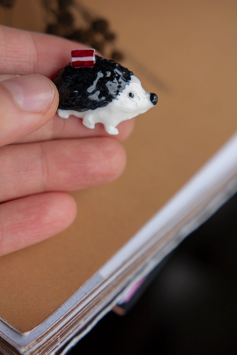 May include: A small, black and white ceramic hedgehog figurine with a red and white striped hat. The figurine is being held in a hand.