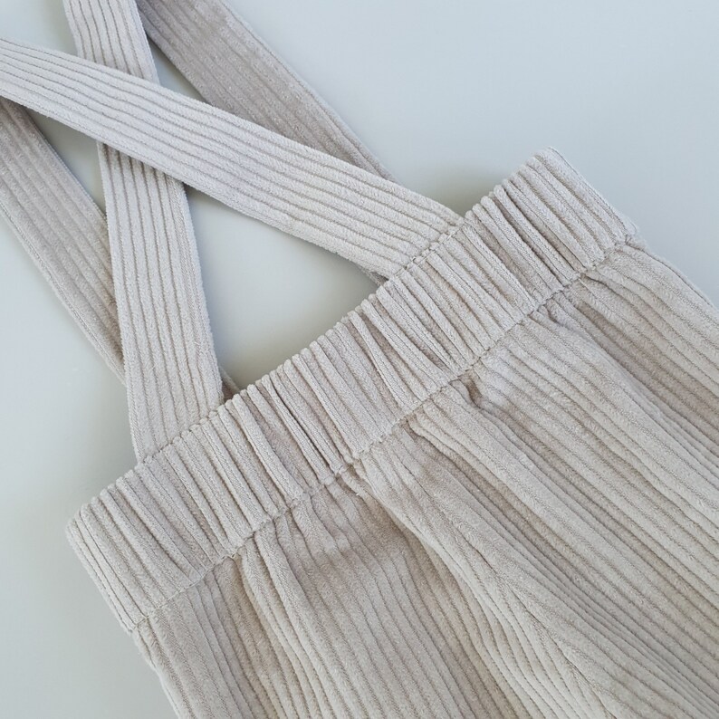 May include: Beige corduroy overalls with adjustable straps. The straps cross in the back and have a wide, comfortable fit.