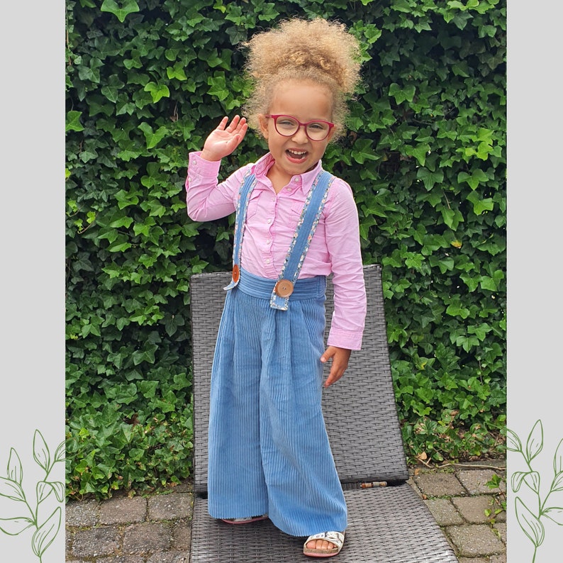 May include: A young girl with curly blonde hair wearing red glasses, a pink button-down shirt, and blue overalls with suspenders. She is smiling and waving with her right hand.