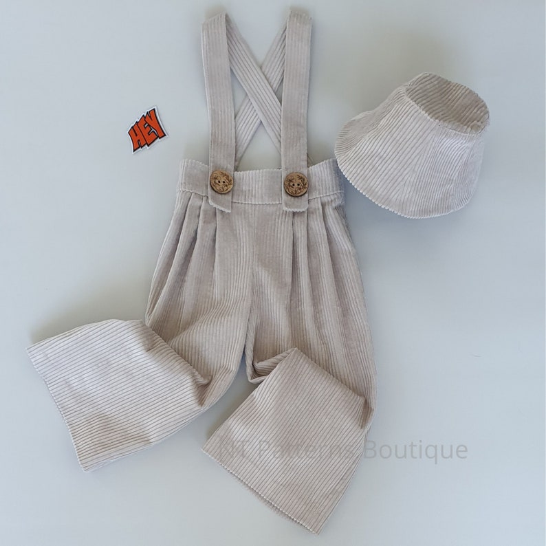 May include: A beige corduroy overall with suspenders and two wooden buttons. The overall has wide legs and comes with a matching beige corduroy bucket hat.