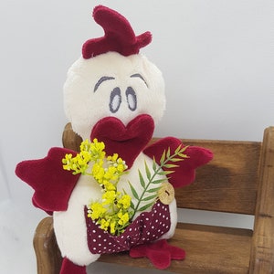 May include: A plush white chicken with red accents sits on a wooden bench. The chicken is holding a bouquet of yellow flowers and a green leaf. The chicken has a red beak and two black eyes. There are small wooden flowers scattered on the floor in front of the bench.