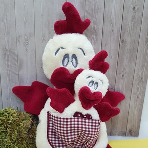 May include: Two plush white chickens with red wings and beaks. The larger chicken has a red and white checkered pocket on its belly. The chickens are sitting on a yellow surface with green and brown speckled eggs and brown twigs.