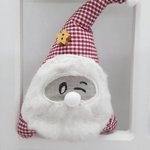 Op de afbeelding: Een rode en witte geruite kerstman-versiering met een witte baard en een knipoogend gezicht. De versiering heeft een stervormige knoop op de muts.