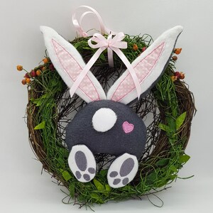 May include: A grey bunny with pink ears and a pink heart on its bottom is attached to a twig wreath with green leaves. The bunny has white feet and a white spot on its bottom. A pink ribbon with a bow is tied around the wreath.