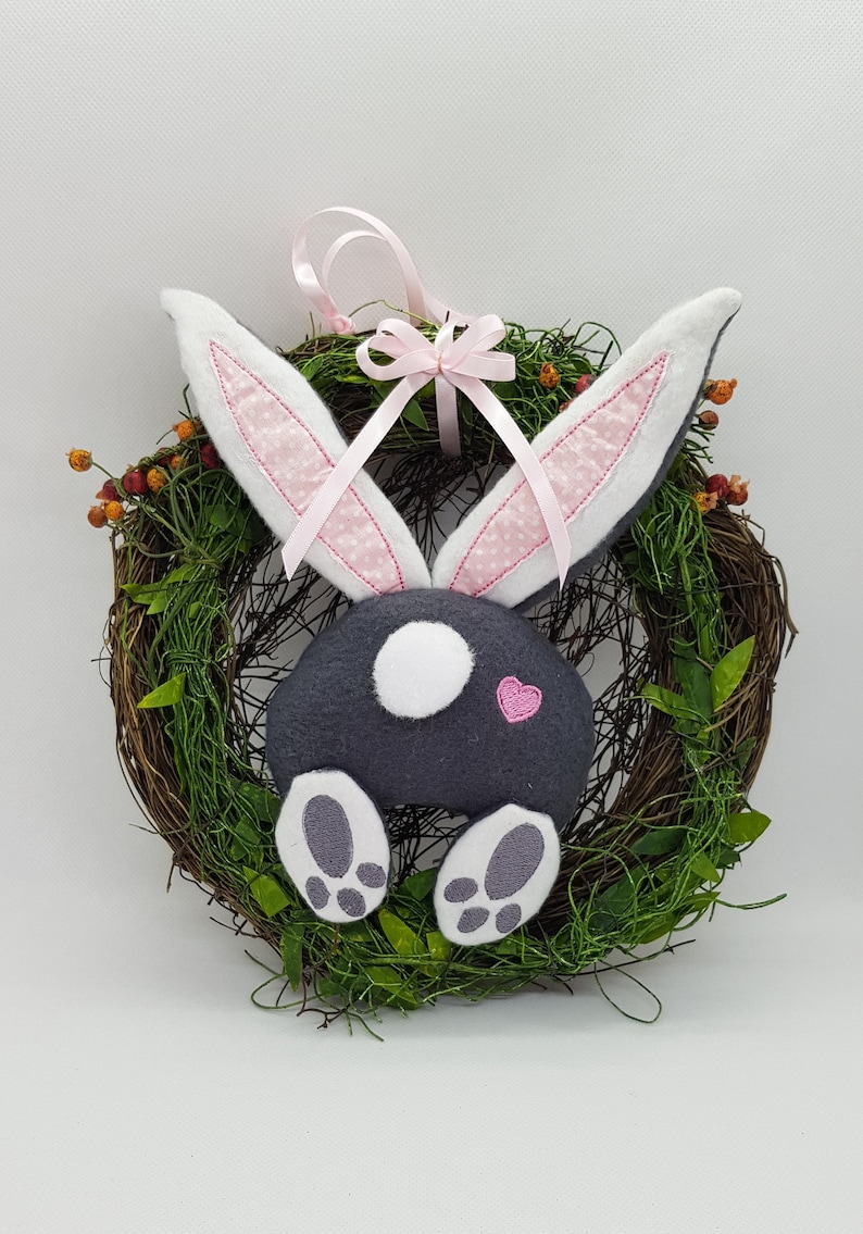 May include: A grey bunny with pink ears and a pink heart on its bottom is attached to a twig wreath with green leaves. The bunny has white feet and a white spot on its bottom. A pink ribbon with a bow is tied around the wreath.