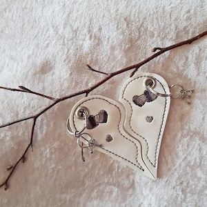 May include: Two white leather keychains shaped like halves of a heart, each with a small silver key ring and a key. The hearts are stitched together and have embroidered gray hearts on them.