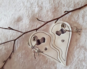 Op de afbeelding: Twee witte leren sleutelhangers in de vorm van twee helften van een hart, elk met een kleine zilveren sleutelring en een sleutel. De harten zijn aan elkaar gestikt en hebben geborduurde grijze harten.