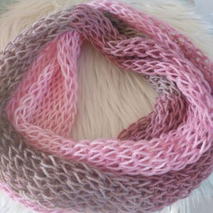 Könnte beinhalten: Handgestrickter Loop-Schal in Rosa- und Brauntönen. Der Schal hat ein offenes Strickmuster, das ihm eine strukturierte Optik verleiht. Die Farben wechseln von einem tiefen Braun zu einem hellen Rosa und erzeugen einen Farbverlauf. Der Schal liegt auf einer weißen, flauschigen Oberfläche.