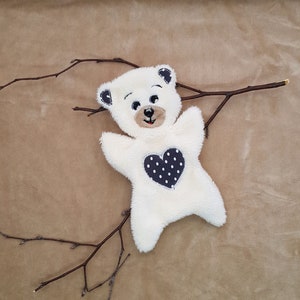 Könnte beinhalten: Weißer Plüschteddybär mit einem schwarz-weißen Herz mit Punkten auf der Brust. Der Bär liegt auf einem braunen Stoffhintergrund.