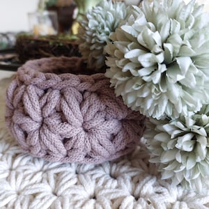 May include: A round, mauve-colored crochet basket featuring a textured star-like stitch pattern. The basket is positioned next to two large, pale green artificial flower heads, with a white crocheted mat visible underneath the basket.