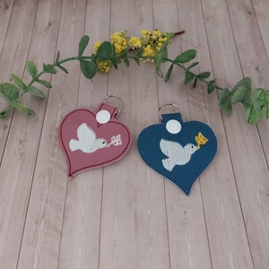 May include: Two heart-shaped keychains with a white dove holding an olive branch. One keychain is pink and the other is blue.