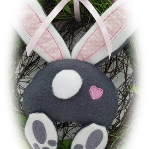 May include: A grey bunny rabbit wreath with pink ears and a pink bow. The bunny has white feet and a white circle on its back. The wreath is made of twigs and green leaves.