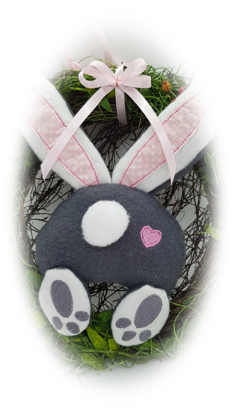 May include: A grey bunny rabbit wreath with pink ears and a pink bow. The bunny has white feet and a white circle on its back. The wreath is made of twigs and green leaves.