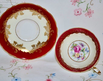 May include: A vintage teacup and saucer set with a red and gold floral design. The cup has a floral pattern in the center and a gold rim. The saucer has a gold floral border around the edge.