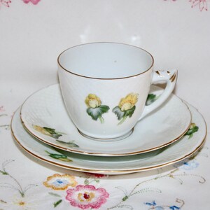 May include: A white teacup, saucer, and side plate set. The set features a gold trim and is decorated with yellow floral designs. The teacup has a curved handle. The set is placed on a floral patterned tablecloth.