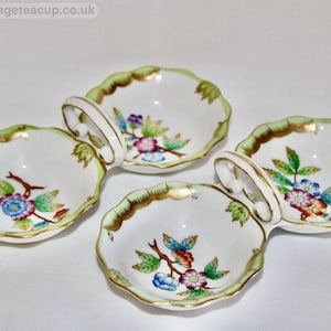May include: Four white porcelain serving dishes with scalloped edges, green trim, and gold accents. Each dish features a handle and a floral design with blue, red, and purple flowers. The dishes are arranged on a white surface.