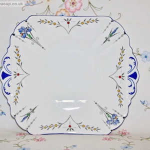 Può includere: Un piatto da torta quadrato bianco con un design floreale blu e oro. Il piatto ha un bordo smerlato ed è decorato con accenti blu, oro e rosso.