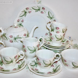 May include: A white porcelain tea set with a strawberry design. The set includes a teapot, creamer, sugar bowl, cups, saucers, and a serving plate. The strawberries are pink with green leaves, and the set is arranged on a white surface.