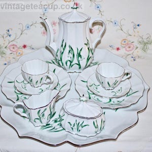 Puede incluir: Juego de té de porcelana blanca con detalles florales verdes, que incluye una tetera, tazas, platillos, una jarra de crema, una azucarera y una bandeja. El juego tiene un borde festoneado y ribete plateado.