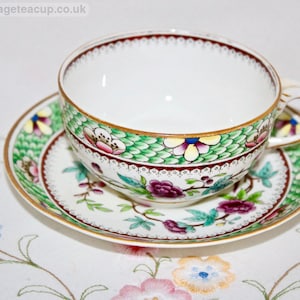 May include: A porcelain teacup and saucer set featuring a white base with a green scale pattern border, gold rim accents, and floral motifs in shades of purple, pink, and green. The text www.thevintageteacup.co.uk is visible in the top left corner.
