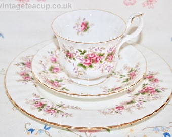 May include: A white teacup and saucer set with a floral pattern of pink roses and green leaves. The set has a gold trim around the edges.