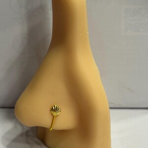 Puede incluir: Un aro de nariz dorado con un pequeño diseño en forma de abanico. La joya está sujeta a un alambre fino y curvado que se engancha en la fosa nasal. El aro de nariz se muestra en una nariz de maniquí.