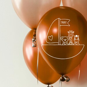 K&ouml;nnte beinhalten: Drei kupferfarbene Ballons mit einem wei&szlig;en Design eines Bootes mit einer Giraffe, einem Zebra und einem B&auml;ren. Der Ballon in der Mitte tr&auml;gt das Wort "Taufe".