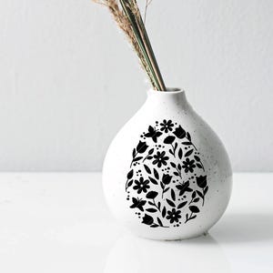 Puede incluir: Jarr&oacute;n de cer&aacute;mica blanco con un dise&ntilde;o floral negro en forma de huevo. El jarr&oacute;n est&aacute; vac&iacute;o y tiene una abertura estrecha en la parte superior.