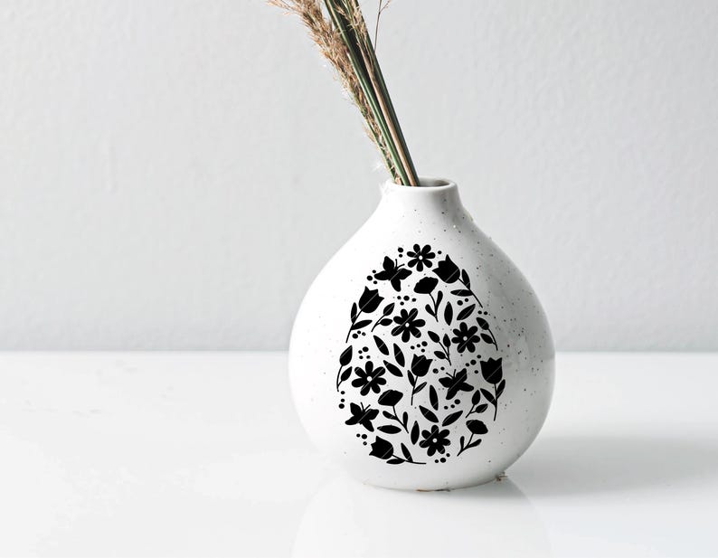 Puede incluir: Jarr&oacute;n de cer&aacute;mica blanco con un dise&ntilde;o floral negro en forma de huevo. El jarr&oacute;n est&aacute; vac&iacute;o y tiene una abertura estrecha en la parte superior.