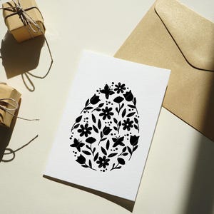 Puede incluir: Una tarjeta blanca con una silueta negra en forma de huevo llena de motivos florales. La tarjeta est&aacute; sobre una superficie blanca con dos regalos envueltos en marr&oacute;n.