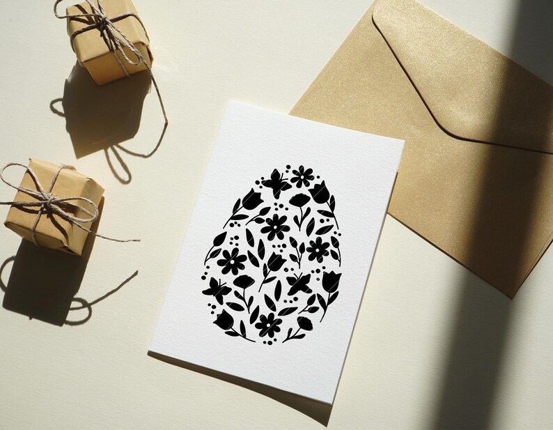 Puede incluir: Una tarjeta blanca con una silueta negra en forma de huevo llena de motivos florales. La tarjeta est&aacute; sobre una superficie blanca con dos regalos envueltos en marr&oacute;n.