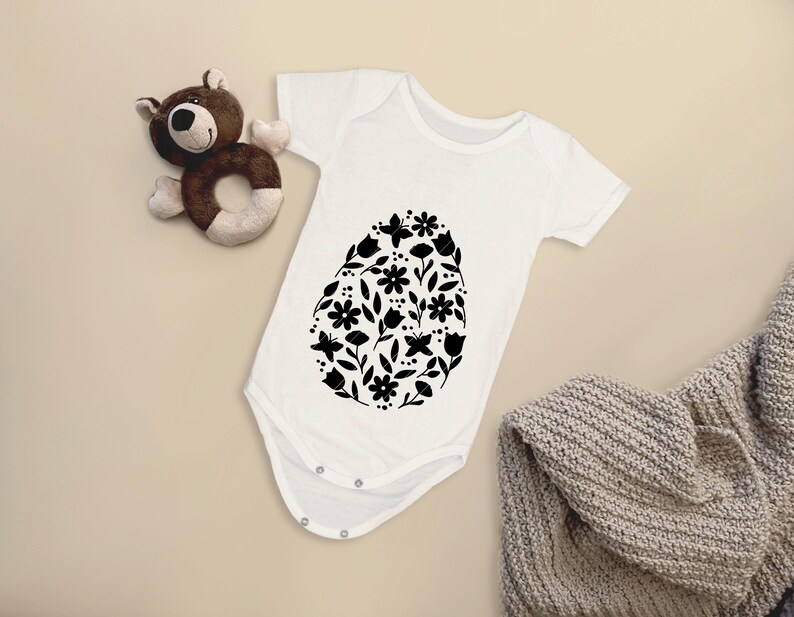 Puede incluir: Un body blanco para beb&eacute; con un dise&ntilde;o floral negro en forma de huevo.