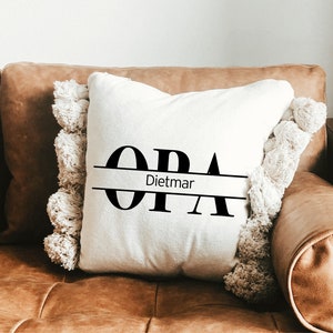 May include: White throw pillow with black text that reads "OPA" with the name "Dietmar" centered below. The pillow has a fringe border and is sitting on a brown leather couch.
