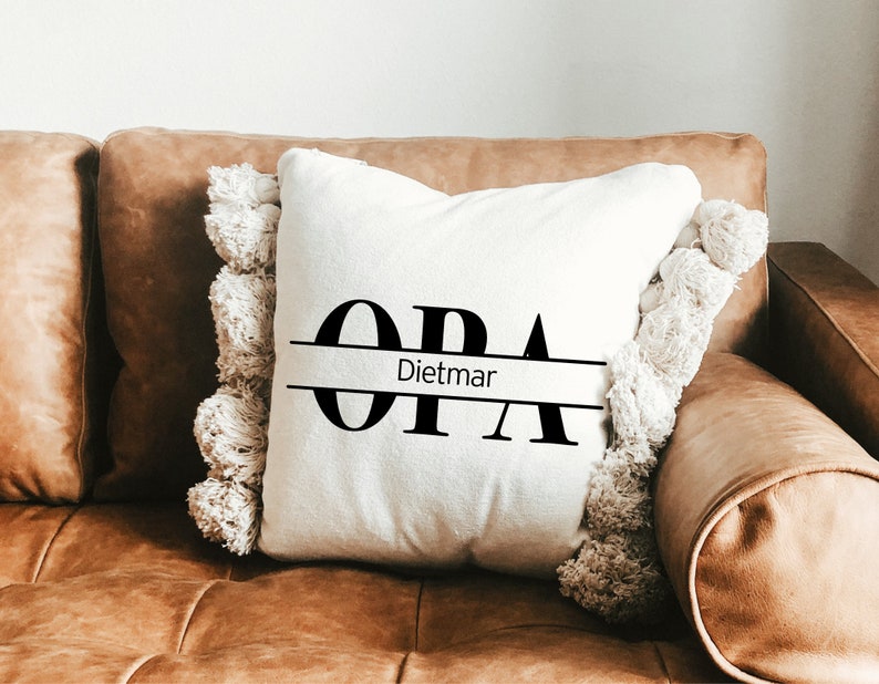May include: White throw pillow with black text that reads "OPA" with the name "Dietmar" centered below. The pillow has a fringe border and is sitting on a brown leather couch.