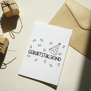 May include: A white card with the text "Geburtstagskind" and a party hat illustration. The card is surrounded by confetti and is on a white surface.