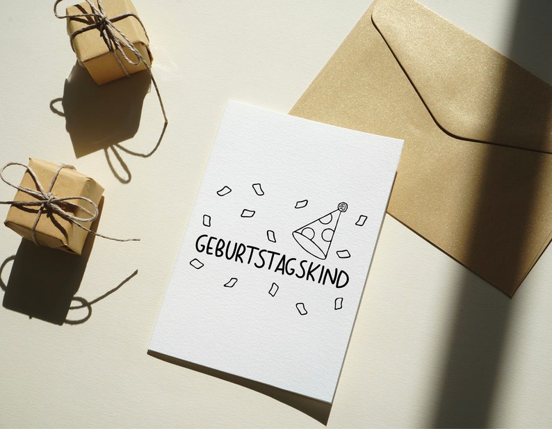 May include: A white card with the text "Geburtstagskind" and a party hat illustration. The card is surrounded by confetti and is on a white surface.