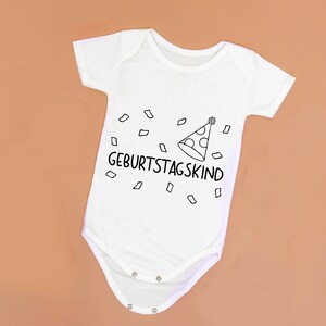 May include: White baby onesie with black text that says "Geburtstagskind" and a party hat with confetti.