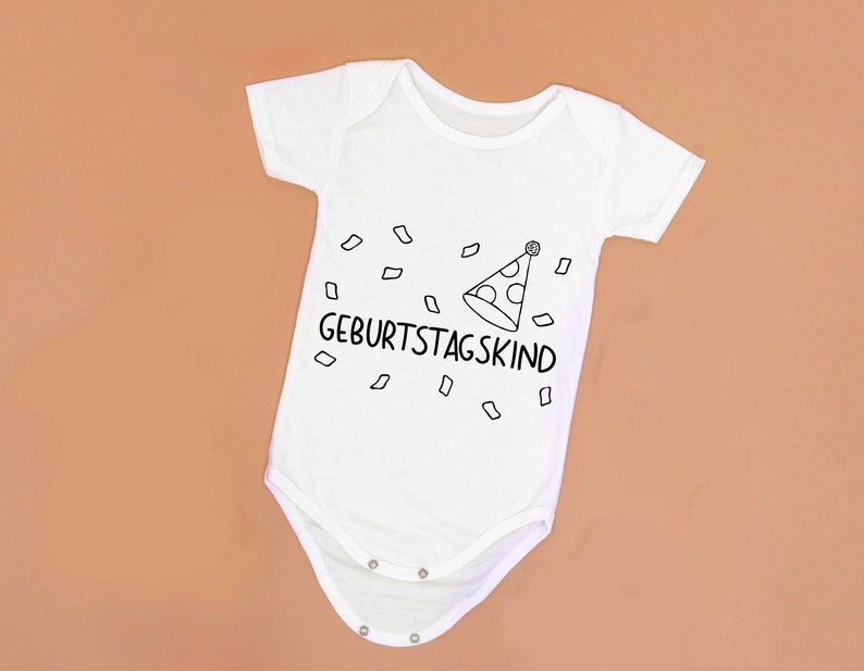 May include: White baby onesie with black text that says "Geburtstagskind" and a party hat with confetti.