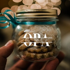 May include: A glass jar with a gold lid and a blue ribbon tied around it. The jar is filled with cookies and has a white label with the text "OPA Nils" on it.