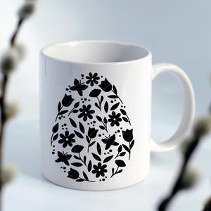 Puede incluir: Taza de cer&aacute;mica blanca con una silueta negra de un dise&ntilde;o en forma de huevo lleno de flores y mariposas.