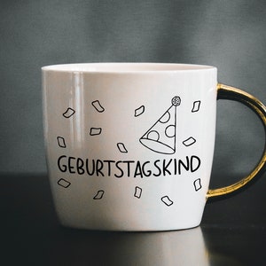 May include: White ceramic mug with a gold handle. The mug has a black and white illustration of a party hat and confetti. The text "Geburtstagskind" is printed on the mug.